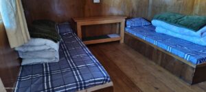 simple wooden room with two single beds, blue and white checkering bedding, folded blankets, a small wooden table, and wooden walls and floors 