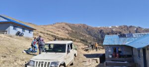 A white vehicle was parked on a dirt road near a few small buildings in a mountainous region, with people standing nearby.
