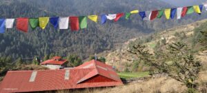Colorful prayer flags strung across a mountainous landscape, with a building with a red roof visible below.