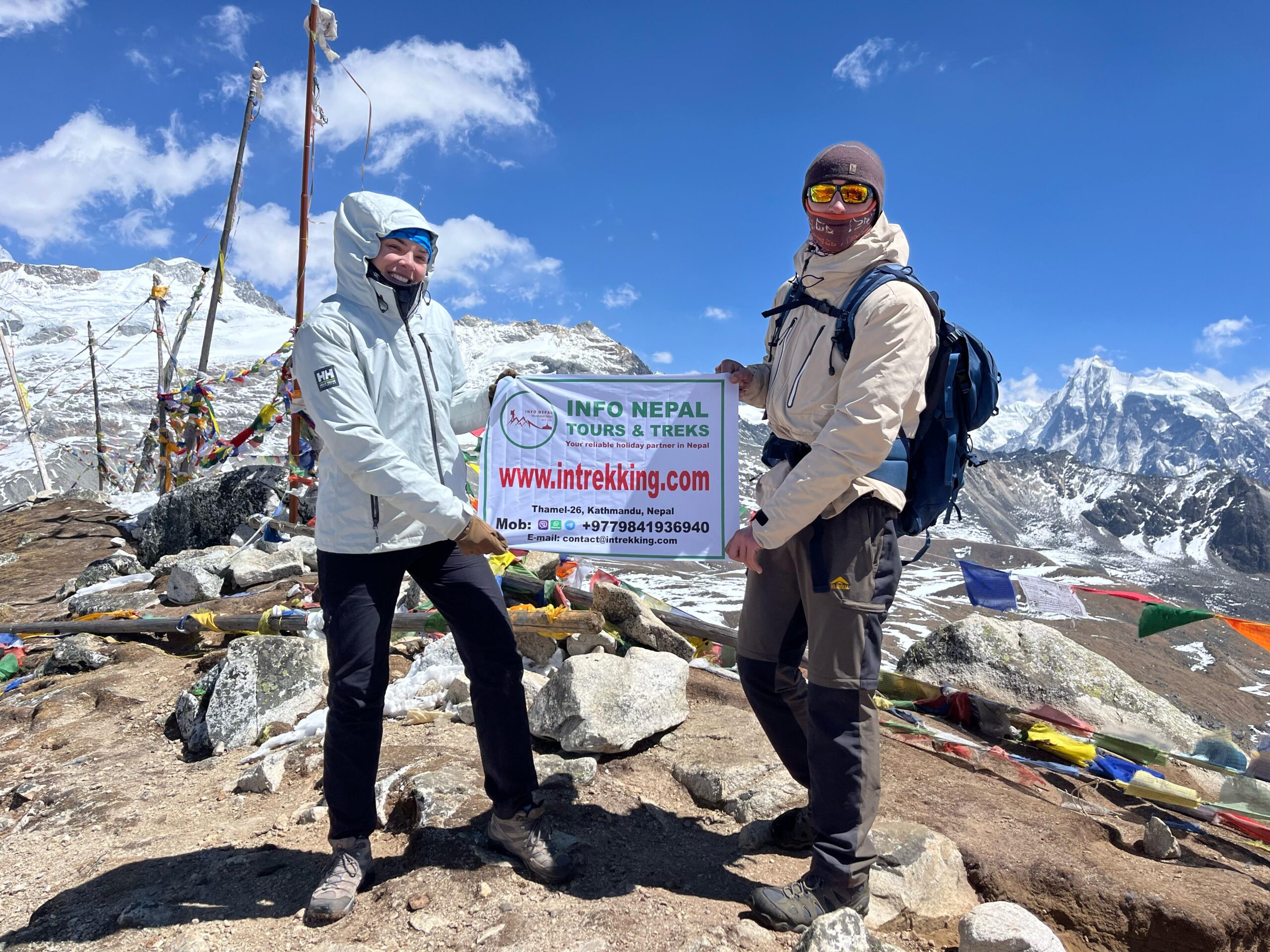 Two trekkers stand on a rocky, mountainous trail, holding a banner for Info Nepal Tours and treks.