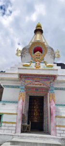 A beautifully adorned white Buddhist stupa with a golden spire, colorful doorway, and golden deer statues under a cloudy sky.