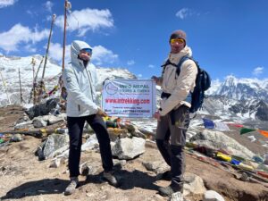 Two trekkers holding banners of info Nepal treks surrounded by mountains and prayer flags behind with snow-capped backdrop 