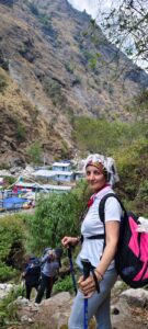 A smiling woman with a white head covering and a pink backpack poses with trekking poles on a rocket trail.