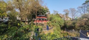 traditional red and white Nepalese building with a from dark roof and porch, surrounded by green trees.